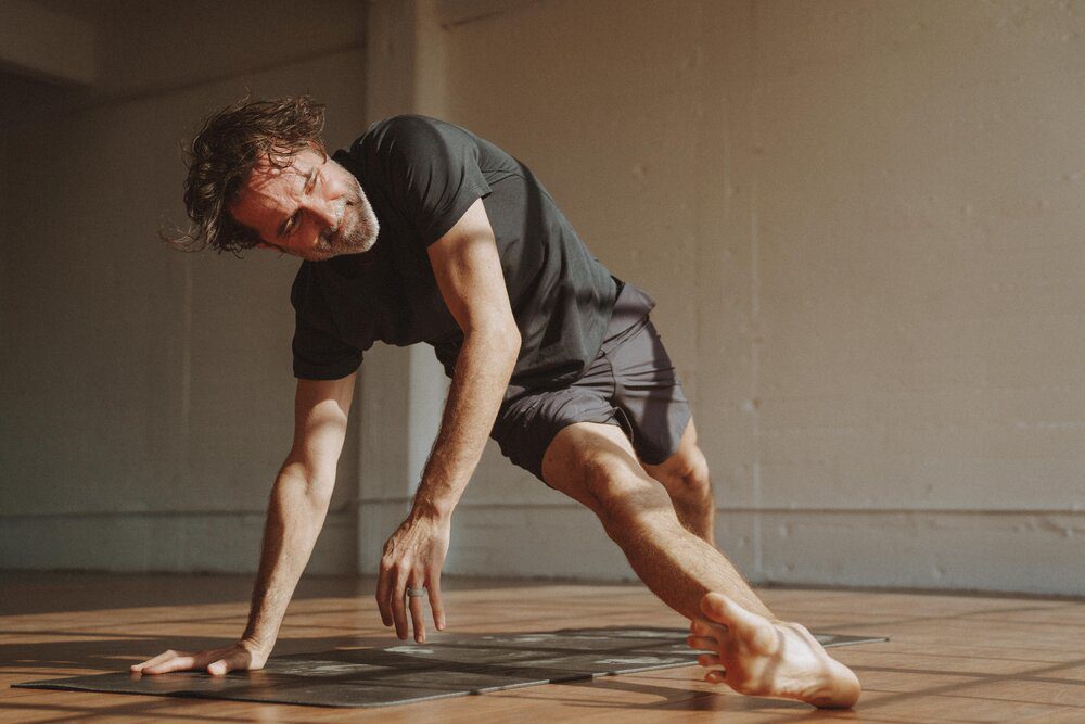 Yoga instructor and The Light Orchard founder Dalton Grant in a black athletic outfit does Fallen Triangle pose (Patita Tarasana) on a yoga mat in a sunlit room.