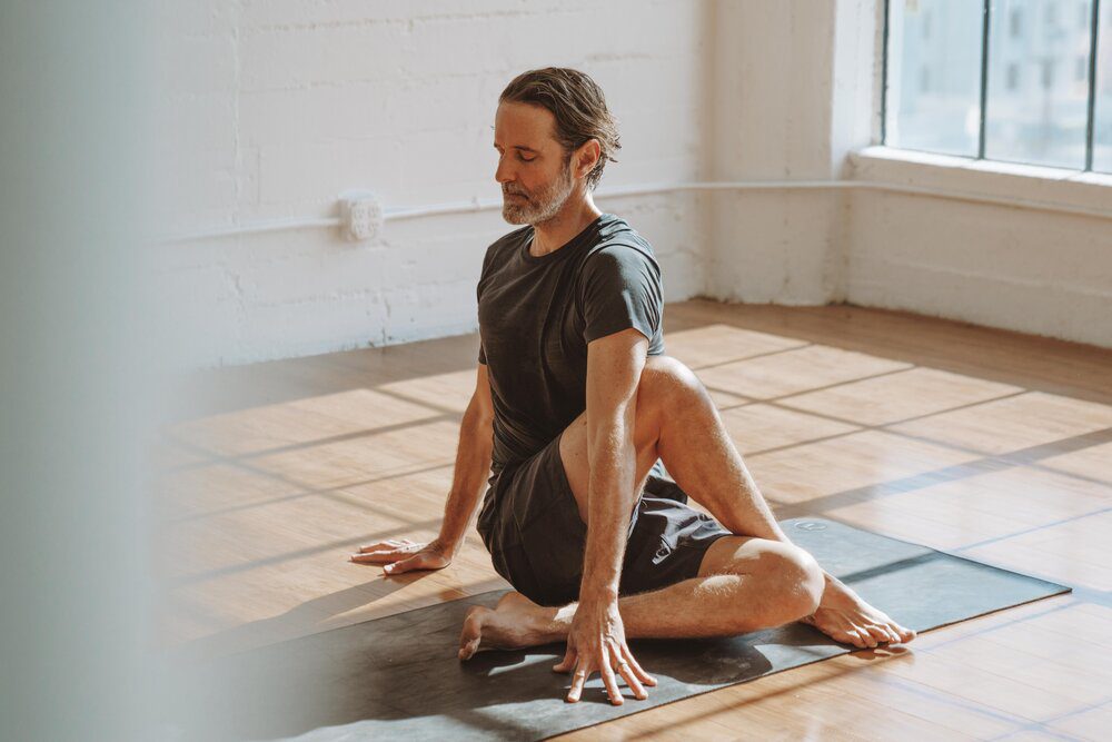 Yoga instructor and The Light Orchard founder Dalton Grant sits on a mat in Lord of the Fishes pose (Matsyendrasana) in a well-lit room, meditating with his eyes closed.
