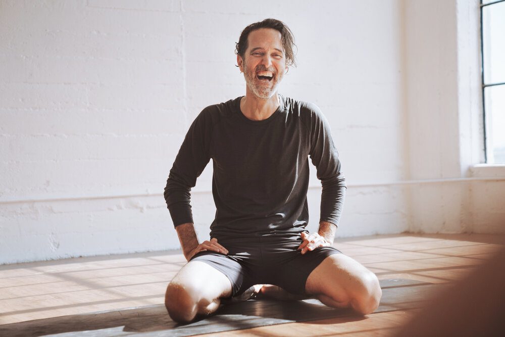 Yoga instructor and The Light Orchard founder Dalton Grant laughs and sits on his heels on a yoga mat in a sunny room.